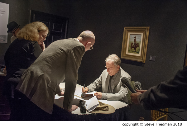 Candid photo of Dick Cavett sitting at a table signing a book with two people standing and leaning over to watch him.